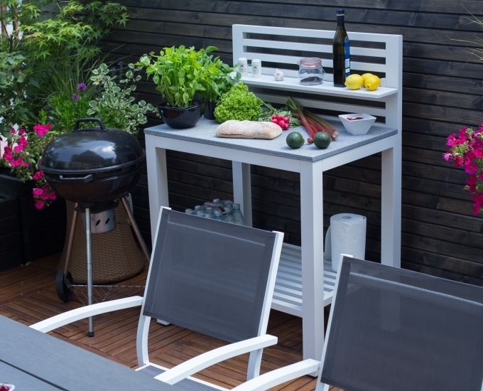 Przytulne patio na świeżym powietrzu z grillem, białym stołem z jedzeniem i napojami, otoczone bujnymi roślinami i kwiatami. dwa szare krzesła stoją naprzeciwko stołu, ustawionego na drewnianym tarasie.