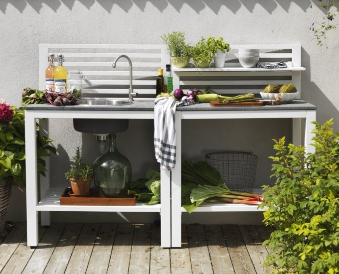 Stanowisko kuchenne na świeżym powietrzu ze zlewem i półkami zaopatrzonymi w świeże warzywa, zioła i przybory kuchenne na słonecznym patio otoczonym zielonymi roślinami.