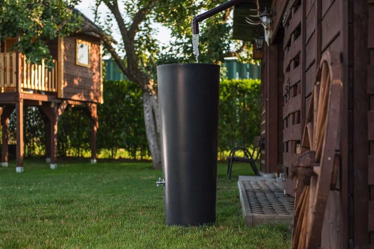 Czarna beczka na deszczówkę zbiera wodę z rury spustowej na dachu obok drewnianej szopy na podwórku. W tle widoczna jest zielona trawa, domek na drzewie i pełen zieleni ogród, tworząc spokojną scenerię na świeżym powietrzu.