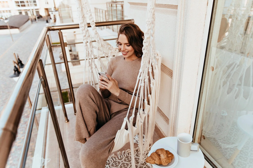 Dziewczyna w przytulnym stroju siedzi na hamakowym krześle na balkonie, uśmiechając się do telefonu. Croissant i kubek leżą na małym stoliku obok niej.