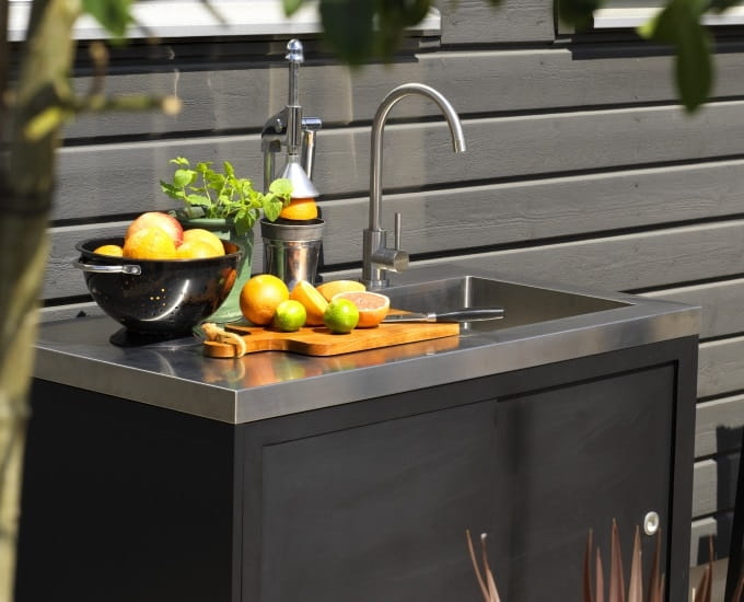 Zestaw kuchenny Coldstream na świeżym powietrzu ze zlewem ze stali nierdzewnej i czarnym blatem z miską owoców cytrusowych, deską do krojenia z cytrynami i rośliną.