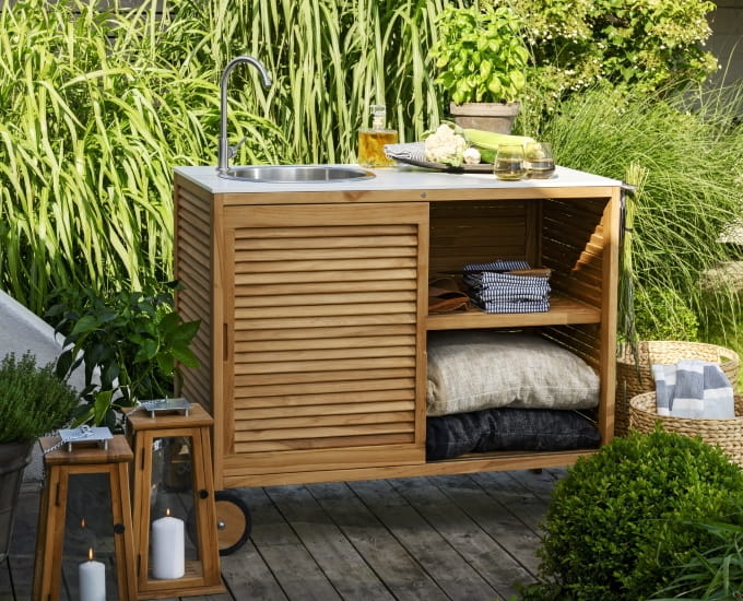 Stylowa drewniana kuchnia ogrodowa Figalia na zewnątrz z wbudowanym zlewem, półkami do przechowywania zastawy stołowej i akcesoriami, na tle bujnej zieleni roślin.