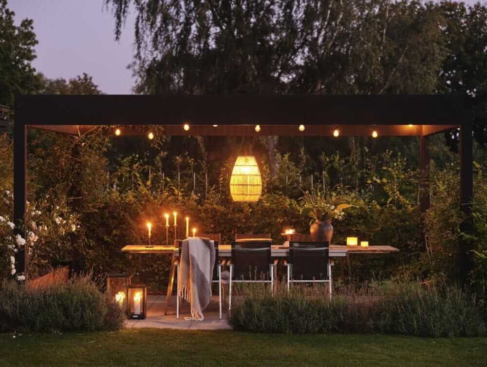 Odkryte patio z drewnianym stołem i krzesłami jest elegancko udekorowane kandelabrami i roślinami doniczkowymi pod pergolą ozdobioną sznurkami świetlnymi i wiszącą latarnią. Przytulną wieczorną atmosferę otacza bujna zieleń.