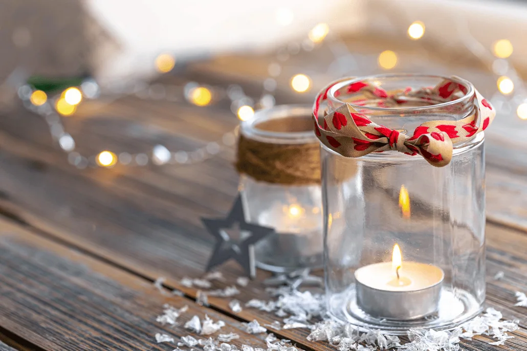 Zapalona świeczka typu tealight w szklanym słoiku z czerwonym szalikiem wokół obręczy stoi na drewnianej powierzchni, z innym słoikiem, srebrną gwiazdą i lampkami sznurkowymi w rozmytym tle.