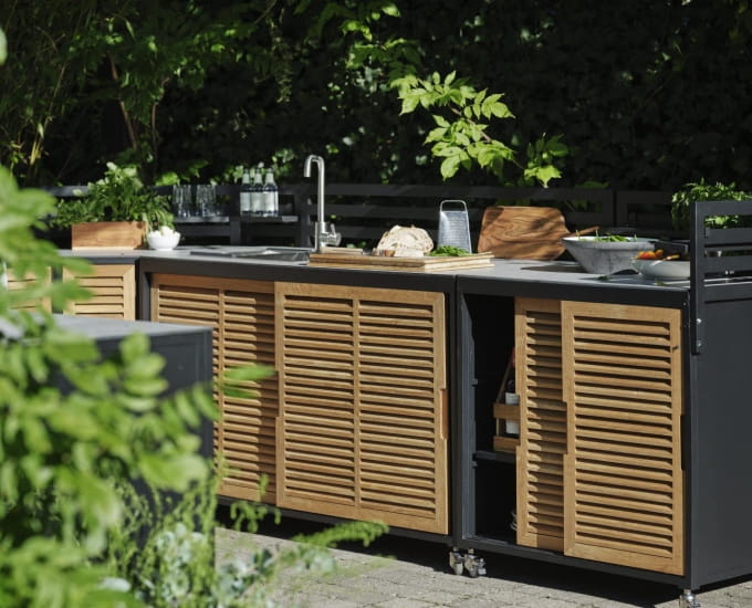 Kuchnia na świeżym powietrzu z drewnianymi szafkami i czarnym blatem, otoczona bujną zielenią w ogrodzie. widoczne elementy obejmują zlew i niektóre przybory kuchenne.