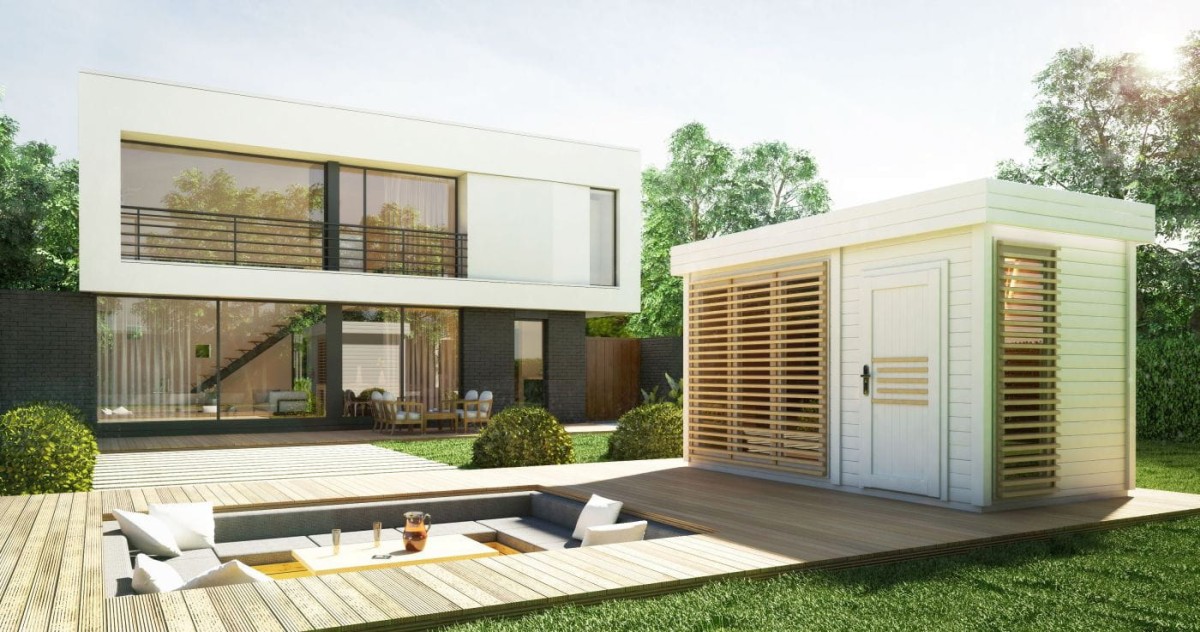 Nowoczesny dwupiętrowy dom z dużymi oknami i płaskim dachem, sąsiadujący z niewielką nowoczesną sauną, z widokiem na część wypoczynkową na drewnianym tarasie, otoczony bujną zielenią.