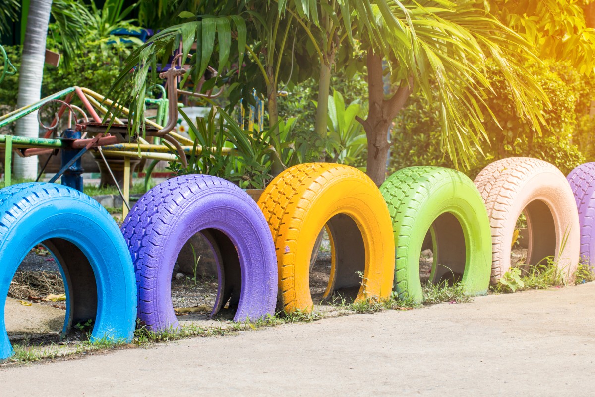Kolorowe, malowane opony ułożone w półkolu i wtopione do połowy w ziemię, wykorzystywane jako zabawowy „Płot z opon” w słonecznym parku z bujną zielenią i urządzeniami zabawowymi