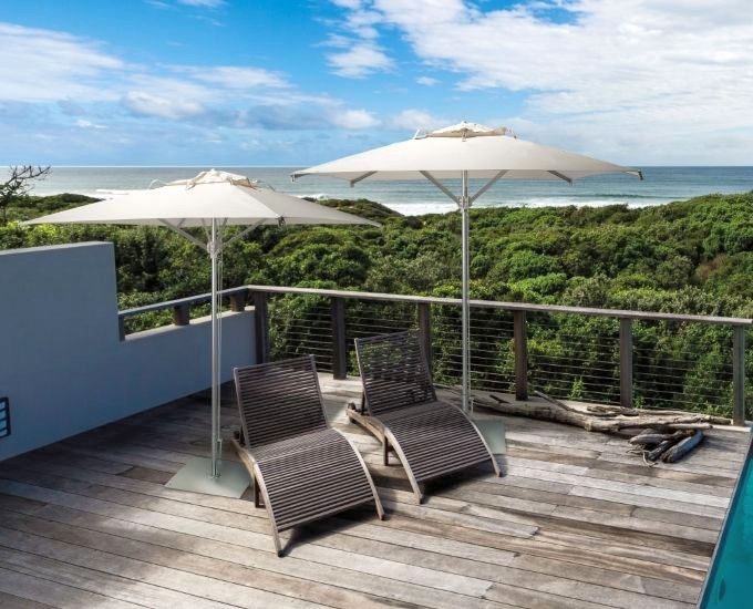 Patio na dachu z dwoma drewnianymi leżakami pod dużymi białymi parasolami wychodzi na bujny zielony las i ocean w tle. Niebo jest częściowo zachmurzone, co dodaje spokoju atmosferze.