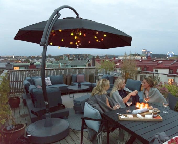 Trzy osoby siedzą na zewnętrznym patio na dachu pod dużym parasolem ozdobionym sznurkami świetlnymi. Są owinięci kocami i rozmawiają przy drinku. W okolicy znajdują się różne układy miejsc do siedzenia, a w tle widać panoramę miasta.