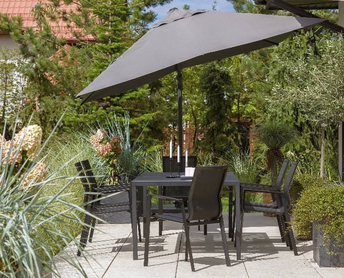 Zestaw na patio na świeżym powietrzu, składający się z czarnego stołu i czterech czarnych krzeseł pod dużym szarym parasolem. Okolica jest pełna zieleni, w tym różnorodnych roślin i kwiatów, tworząc spokojną, ogrodową atmosferę. Wiszące girlandy świetlne dodają nastroju.