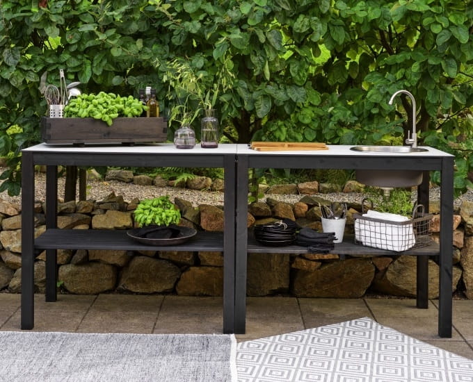 Zestaw kuchenny na świeżym powietrzu z czarnym stołem z wbudowanym zlewem, otoczony bujną zielenią i kamienną ścianą. stół ozdobiony jest talerzami, przyborami kuchennymi i koszem świeżych warzyw.