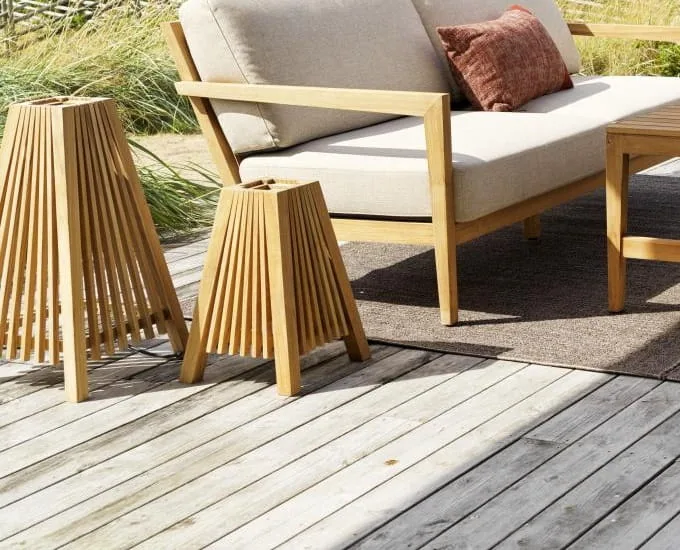 Drewniane meble ogrodowe na tarasie obejmują beżową wyściełaną sofę z brązową poduszką i pasujący stolik. W pobliżu stoją dwie drewniane latarnie z listew, a w tle widać wysoką trawę, co tworzy spokojną scenerię. 