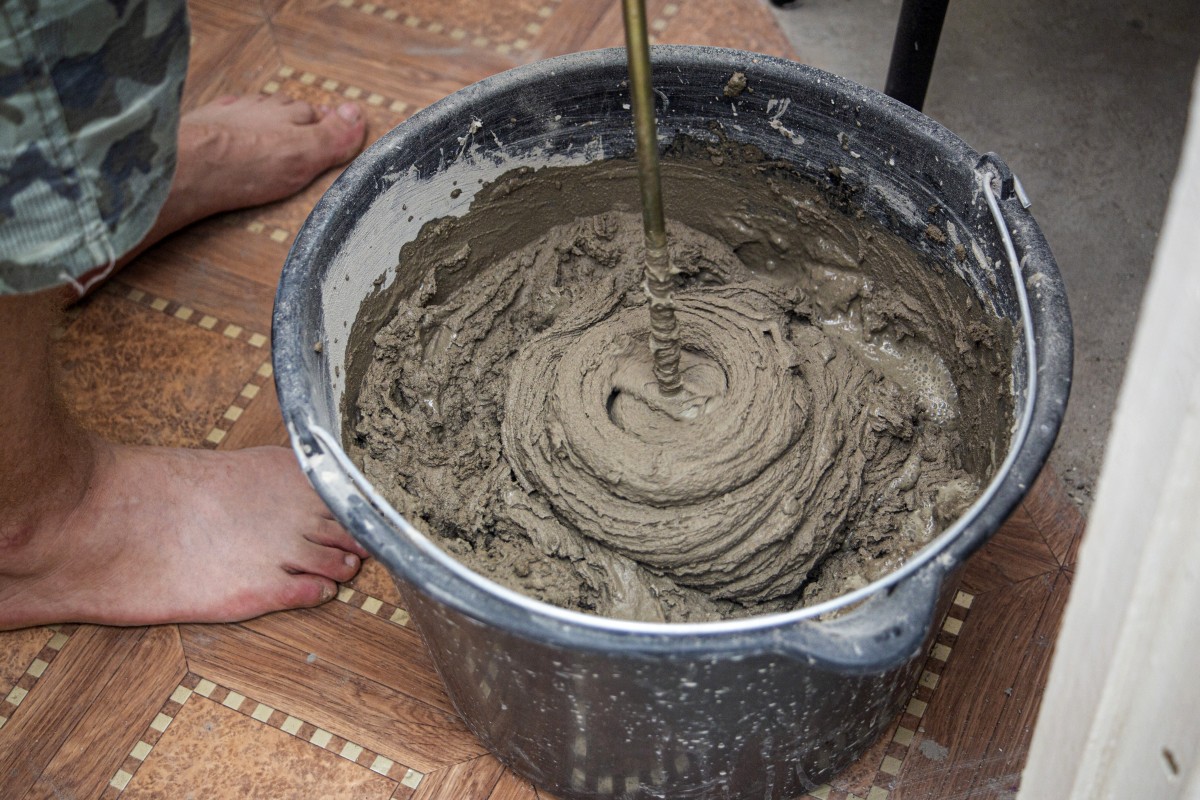 Zdjęcie przedstawiające zbliżenie wiadra wypełnionego świeżo wymieszanym, mokrym cementem. W pobliżu wiadra widać bosą stopę człowieka, a metalowe narzędzie miesza mieszaninę, tworząc spiralę