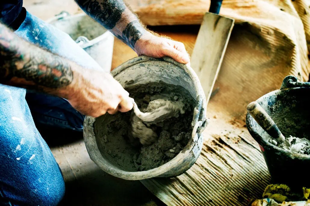 Osoba z wytatuowanymi ramionami miesza cement w wiadrze za pomocą kielni. W pobliżu znajdują się narzędzia i materiały budowlane, w tym kolejne wiadro. 