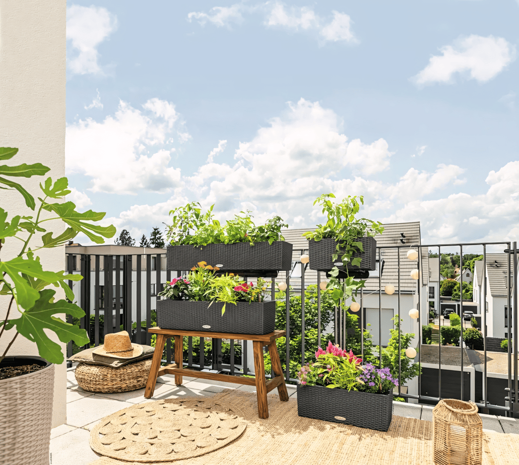 Słoneczny balkon z plecionymi donicami wypełnionymi różnymi zielonymi roślinami i kolorowymi kwiatami, drewniana ławka, okrągły jutowy dywanik, koszyk, słomkowy kapelusz i domy w tle pod błękitnym niebem z chmurami.